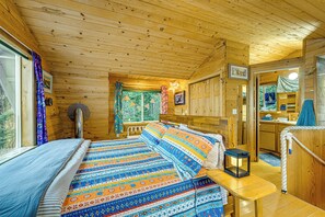 A wooden cabin with a bed, a table with a lantern, a window with curtains, and a bathroom visible through an open door.