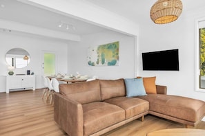A modern living room with a brown leather sofa, a dining area with a white table, and a mounted flat-screen TV.