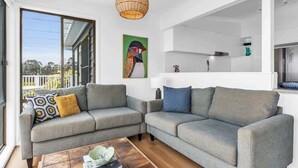 A modern living room with a grey sectional sofa, a coffee table, and a large window with a view of greenery. - Pambula Little Beach House (Pambula Beach)