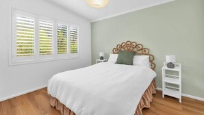 A bedroom with a bed, a window with blinds, a bedside table with a lamp, and a wall-mounted light fixture. - Pambula Little Beach House (Pambula Beach)
