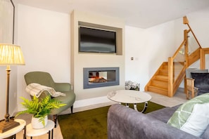 A living room with a green armchair, a grey sofa, a small round table, a floor lamp, a potted plant, a fireplace, and a wooden staircase.