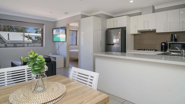 A modern kitchen with white cabinetry, a built-in oven, and a microwave. There's a dining area with a wooden table and chairs, and a TV mounted on the wall. - Sunny 2-Bed in Quiet Location with Balcony (Cannington)