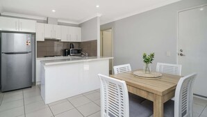 A modern kitchen with white cabinets, a stainless steel refrigerator, a microwave, and a coffee maker. There is a wooden dining table with white chairs and a vase of green plants. - Sunny 2-Bed in Quiet Location with Balcony (Cannington)