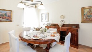 A dining room with a round wooden table, white chairs, and a chandelier. There are paintings on the walls and a wooden cabinet with decorative items.