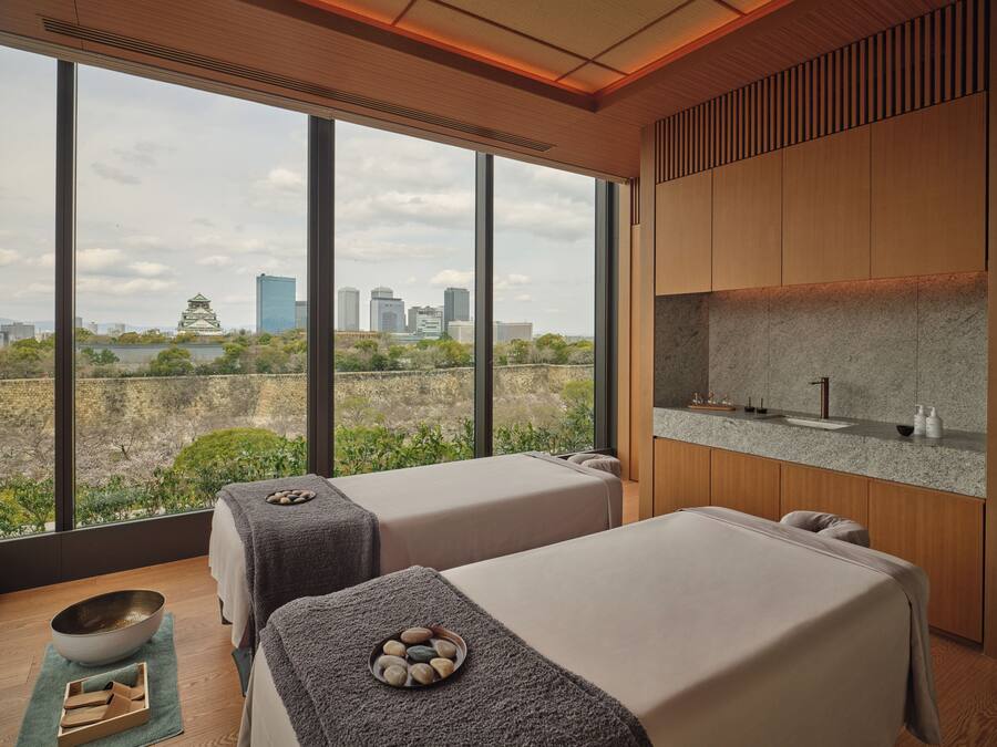 Ein moderner Wellnessraum mit zwei Massagetischen, einer Steinsinnette und Blick auf die Stadtlandschaft.