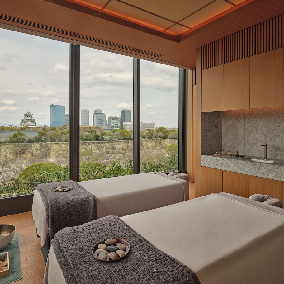 A modern spa room with two massage tables, a stone basin, and a view of a cityscape.