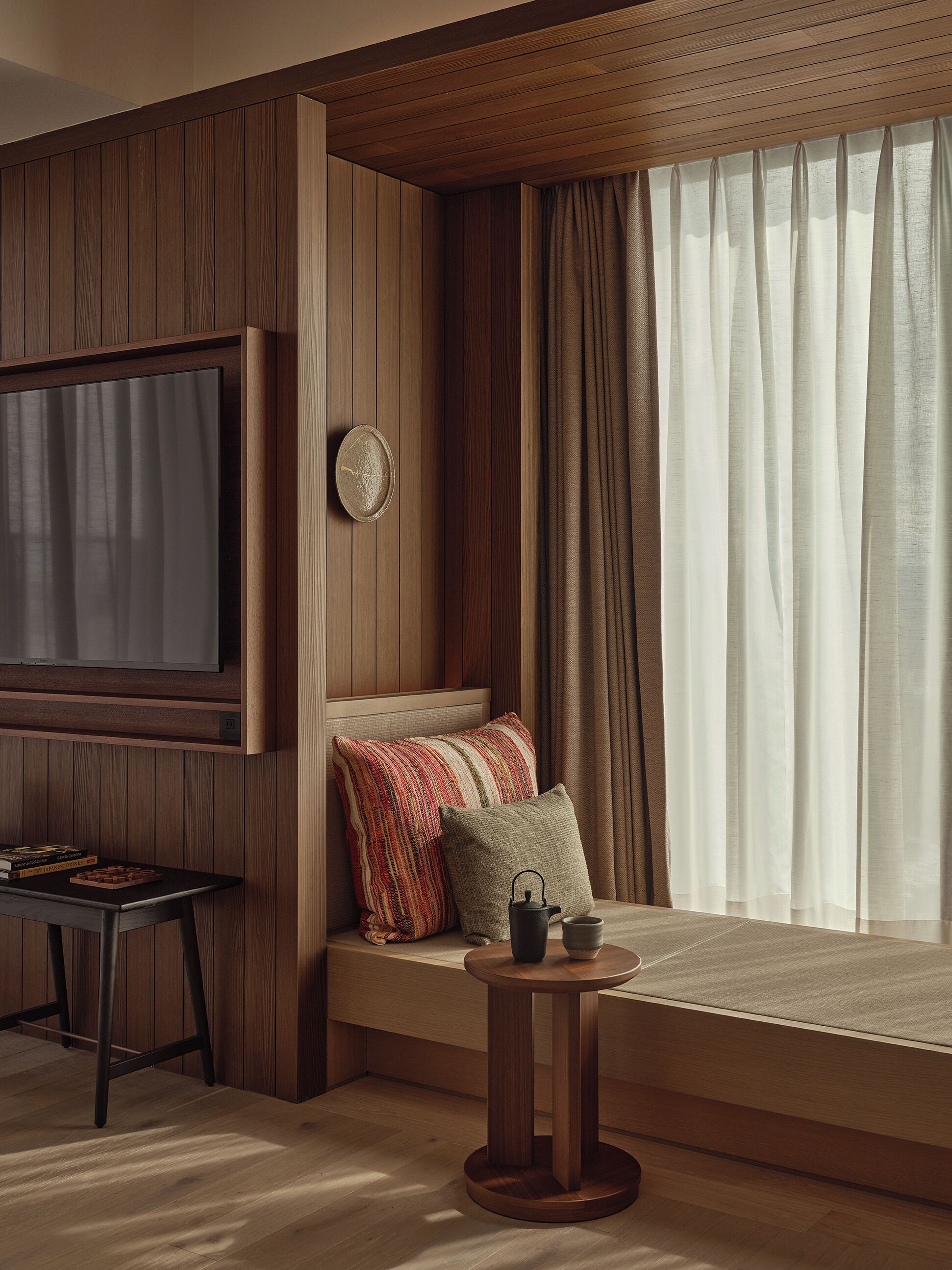 Una habitación acogedora con pared de paneles de madera, un televisor de pantalla plana grande, una zona de estar cómoda con cojines, una pequeña mesa redonda con una tetera y una ventana con cortinas.