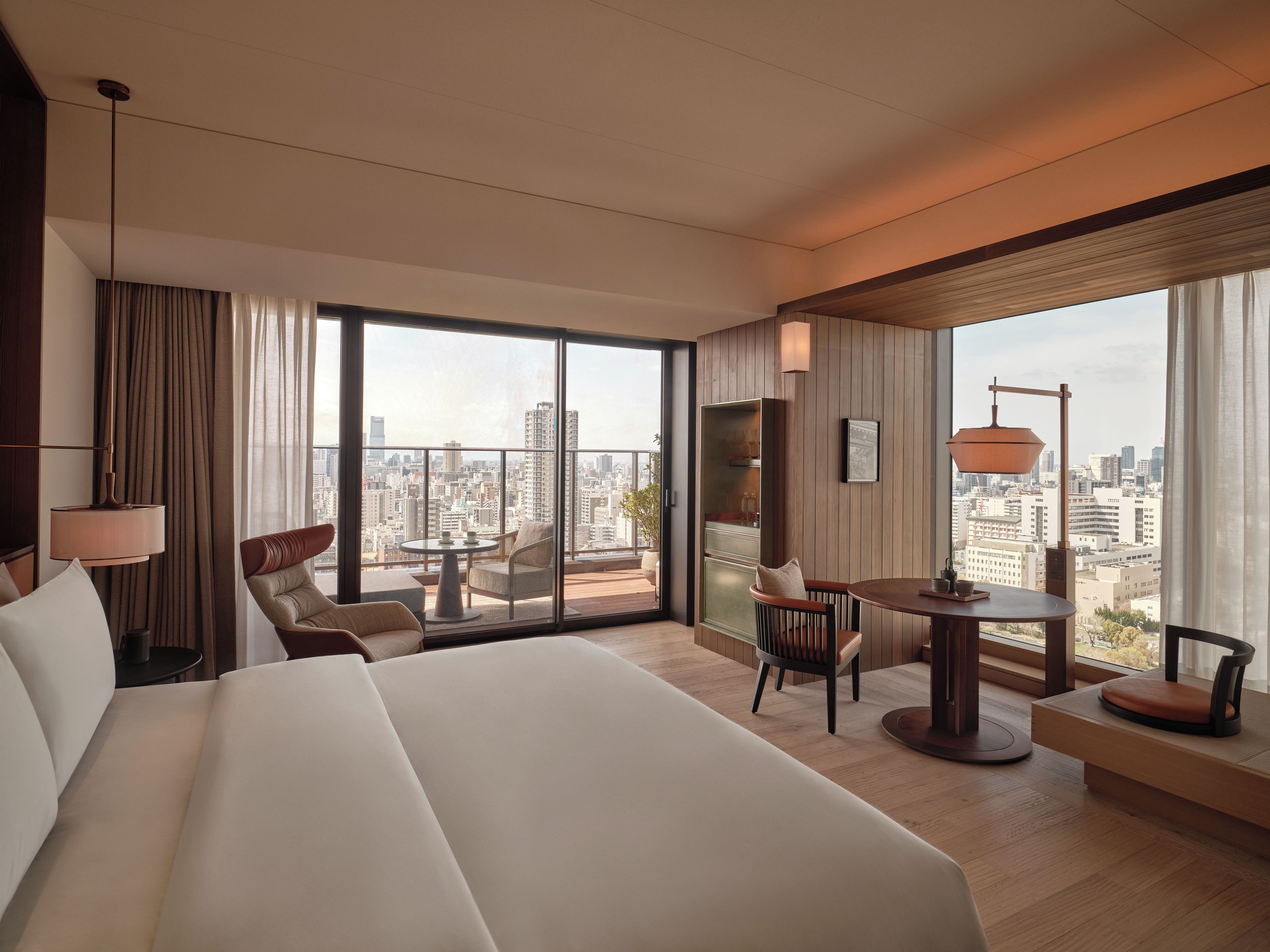 A hotel room with a large bed, a seating area with a table, and a city view through large windows.