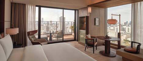 A hotel room with a large bed, a seating area with a table, and a city view through large windows.
