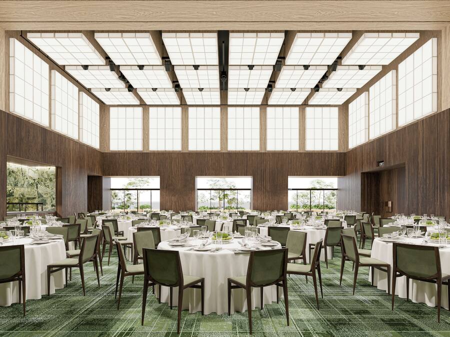 Un amplio salón de banquetes con varias mesas redondas dispuestas para comer, grandes ventanales que permiten la entrada de luz natural, y un techo alto con estructura de celosía.
