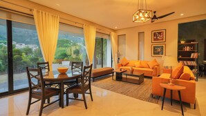 A modern living room with a dining area, large windows with curtains, a sofa set, and a bookshelf.