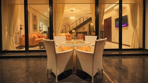 A modern interior with a glass dining table surrounded by wicker chairs, a staircase, and a living area visible through large glass doors.