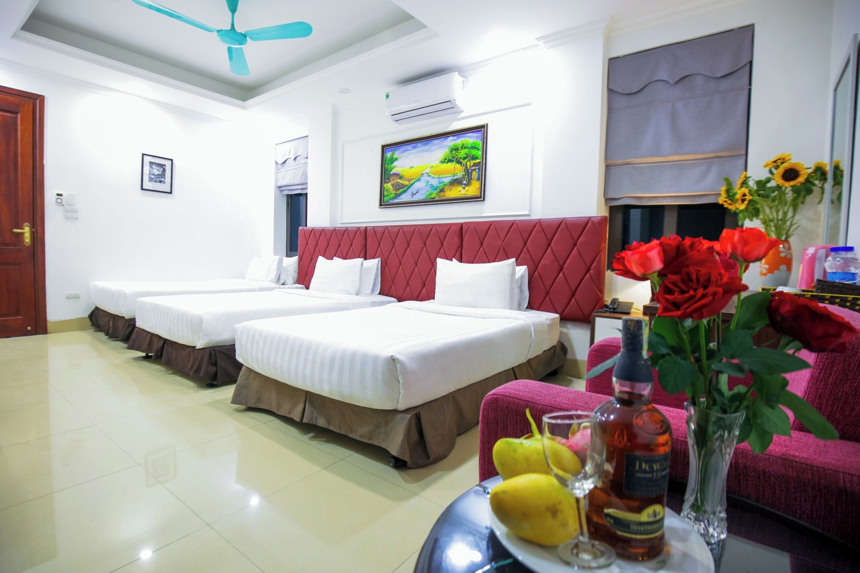 A hotel room with two beds, a red headboard, a painting on the wall, a sofa, and a table with a vase of flowers and a bottle.
