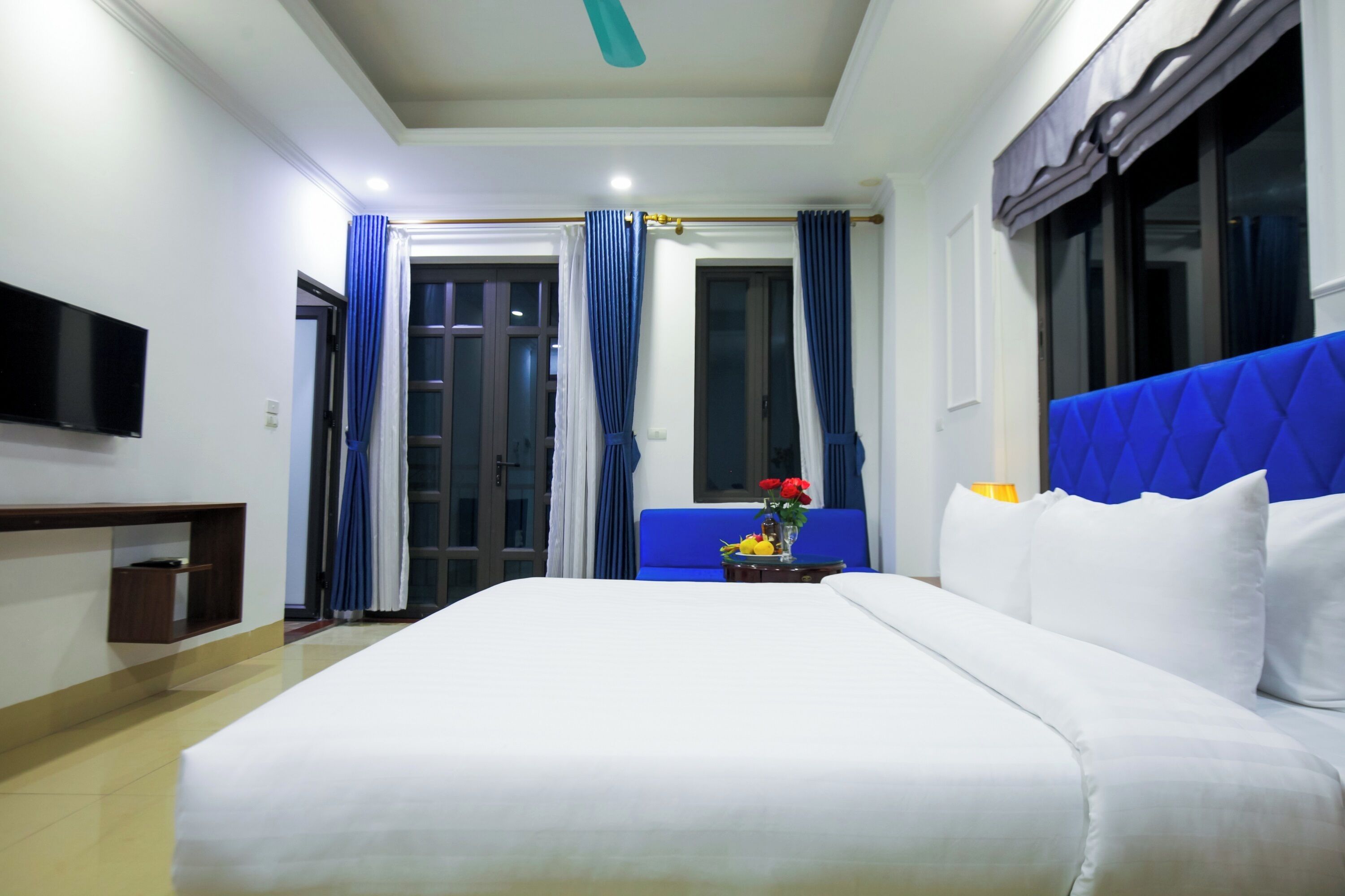 A hotel room with a large bed, a flat-screen TV mounted on the wall, a small table with a vase of flowers, and a window with blue curtains.