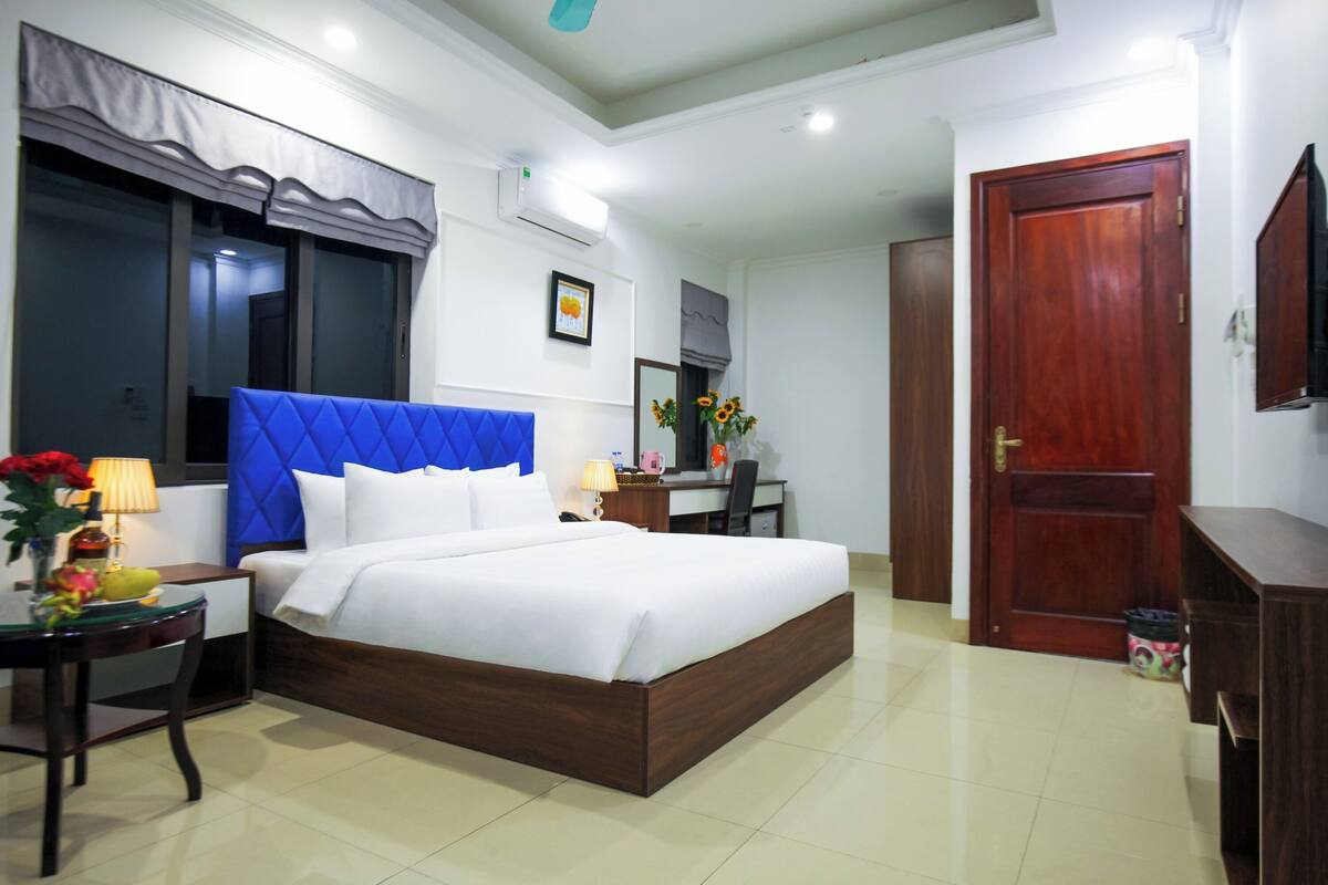 A hotel room with a large bed, a desk, a chair, a small table with a lamp, and a door.