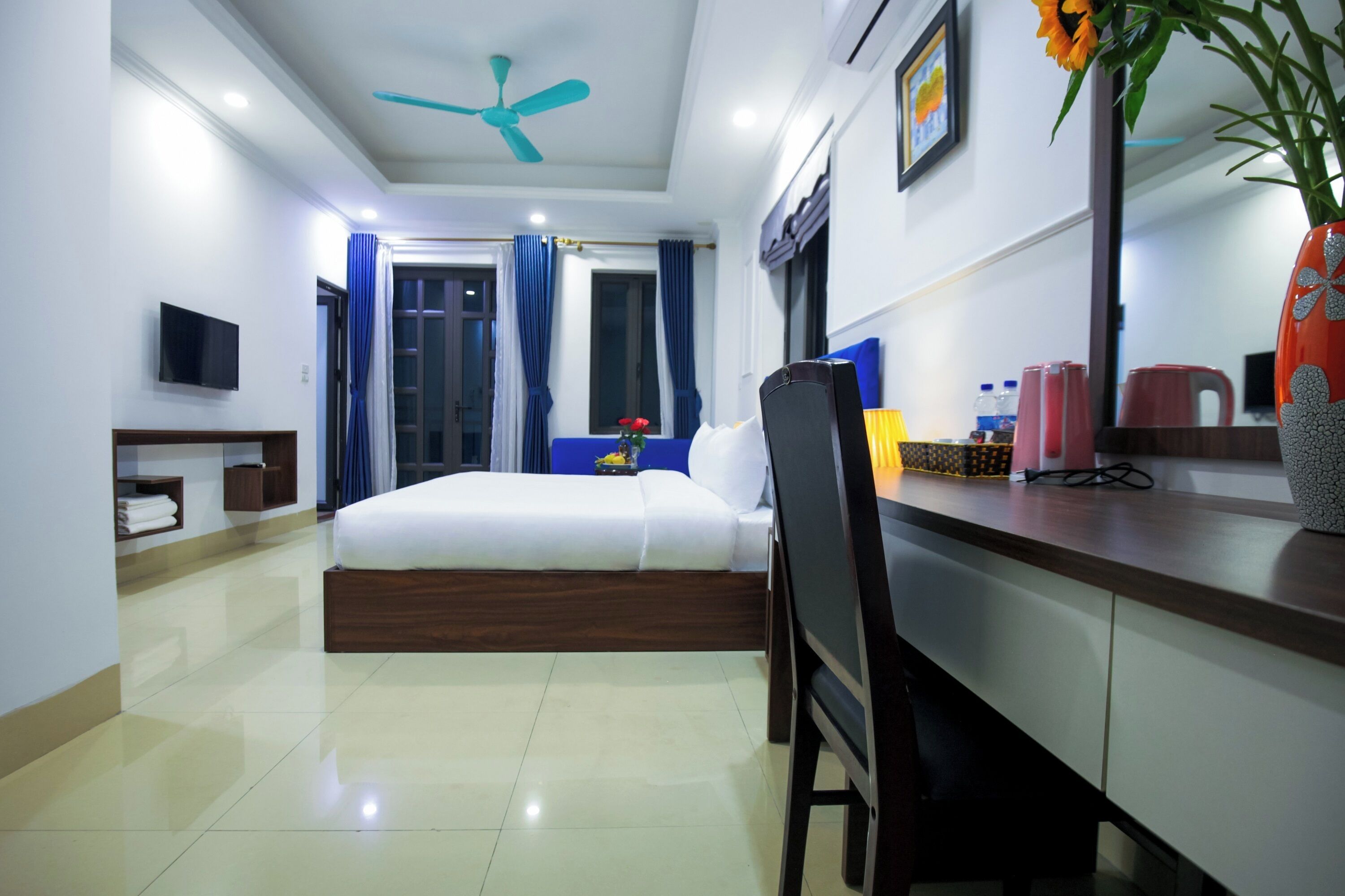 Habitación de hotel con cama, escritorio, silla, televisor, ventilador de techo y un florero con flores.