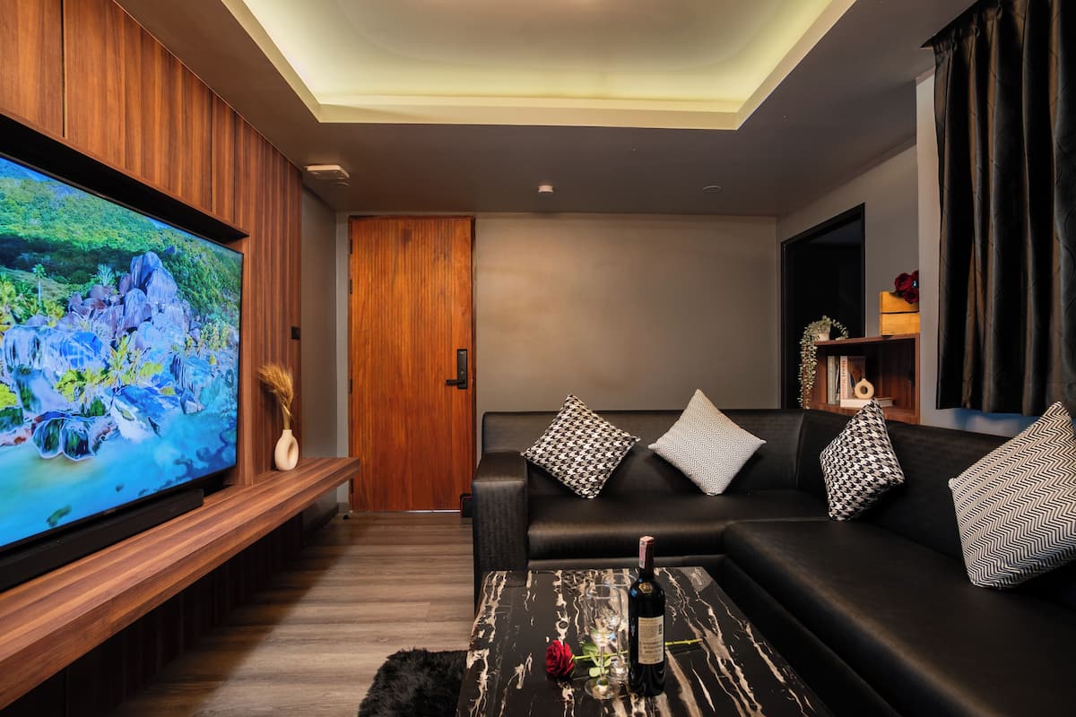 A modern living room with a large flat-screen TV, a black leather sofa with patterned cushions, a wooden entertainment unit, and a marble coffee table with a bottle and glass.
