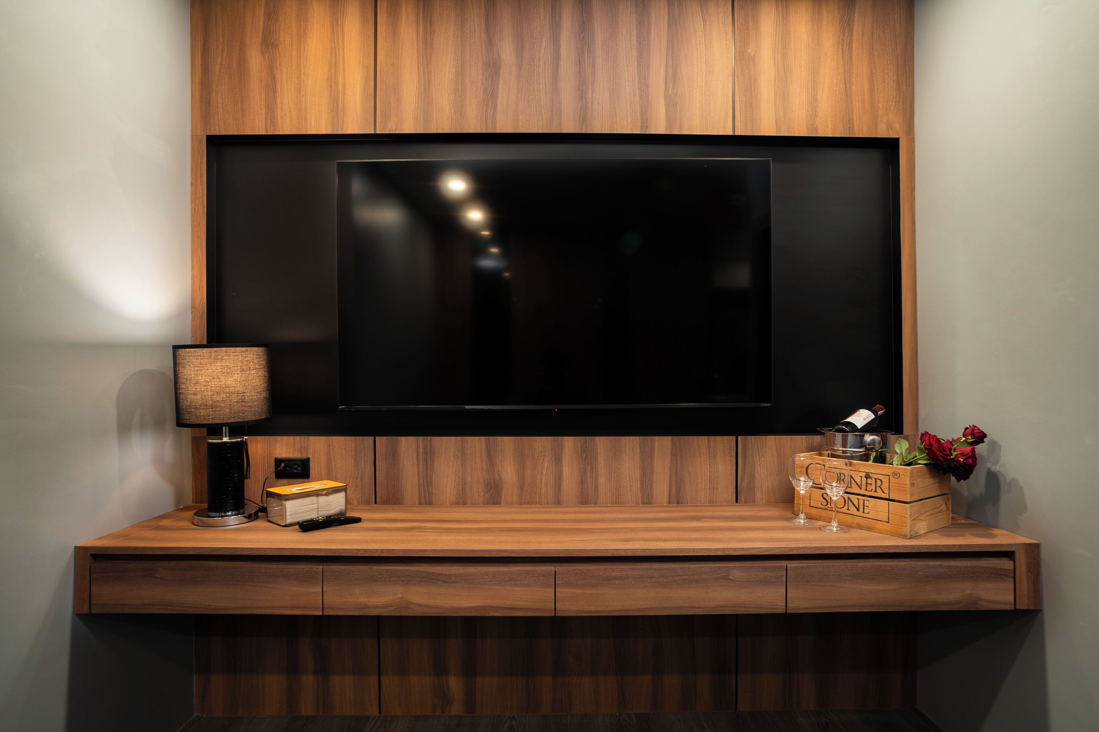 Et moderne tv-møbel med et fladskærms-tv, en lampe, en fjernbetjening og en trækasse med en vinflaske og blomster.