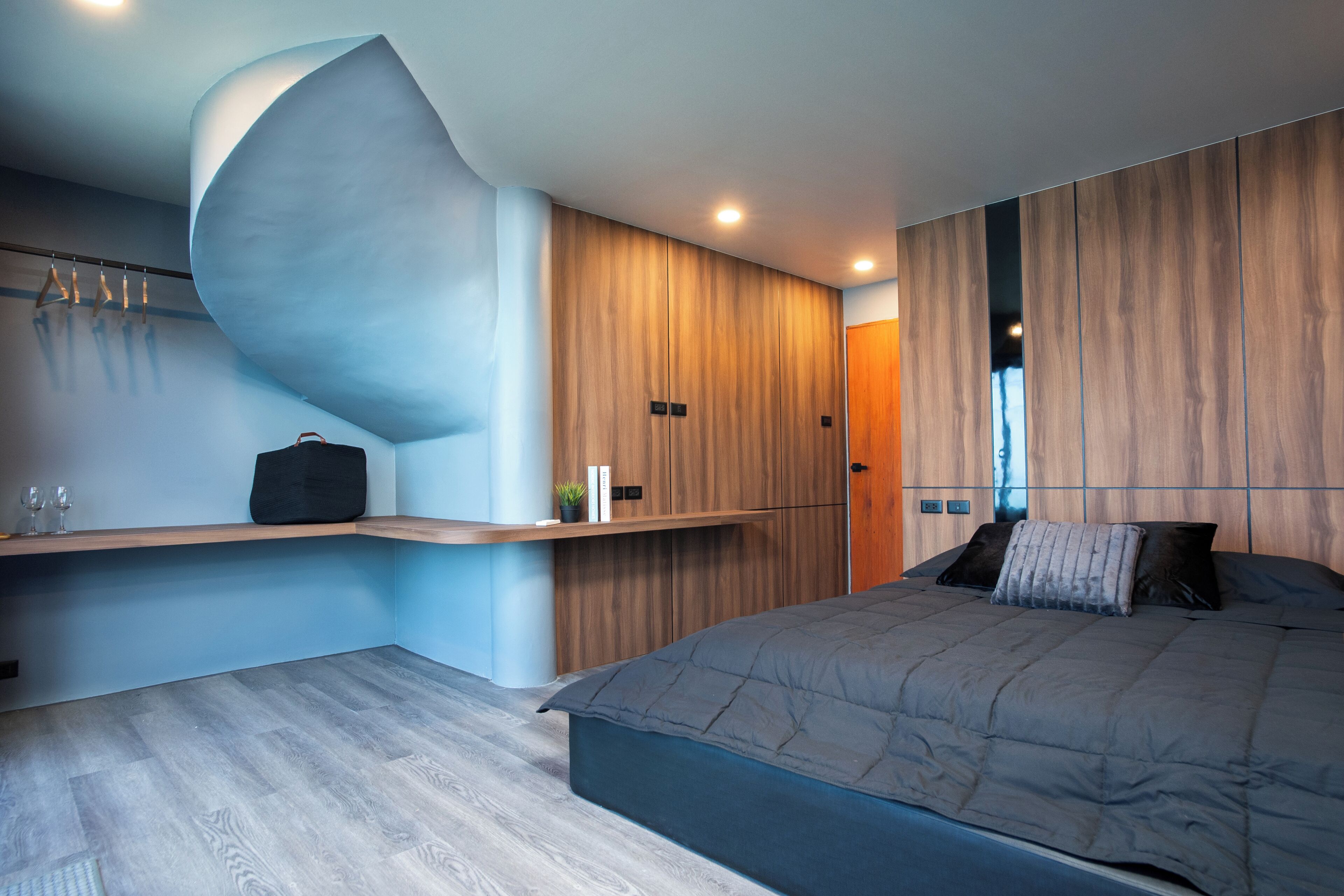 Un dormitorio moderno con una pared curva, una cama con cubrecama gris, paneles de madera y un banco empotrado con un bolso negro y libros.
