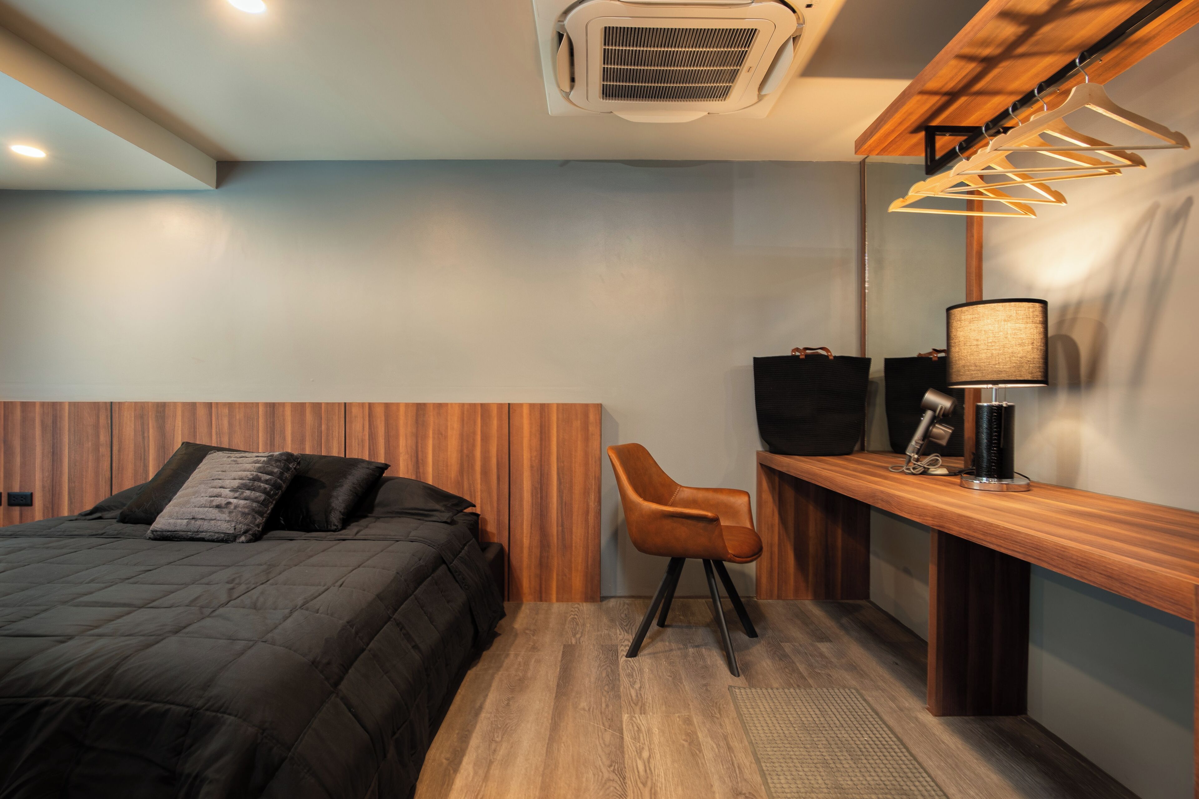Een moderne hotelkamer met een houten hoofdbord, een bed met donkere beddengoed, een houten bureau met een stoel, een lamp en een kledingrek.