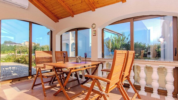 A wooden table and chairs on a balcony with large windows, a clock, and a view of greenery and buildings. - Holiday home Suric (Privlaka)