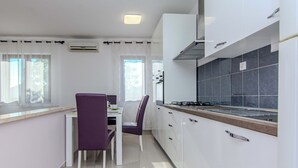 A modern kitchen with white cabinets, a gas stove, and a dining area with a purple chair and white table. - Holiday home Suric (Privlaka)