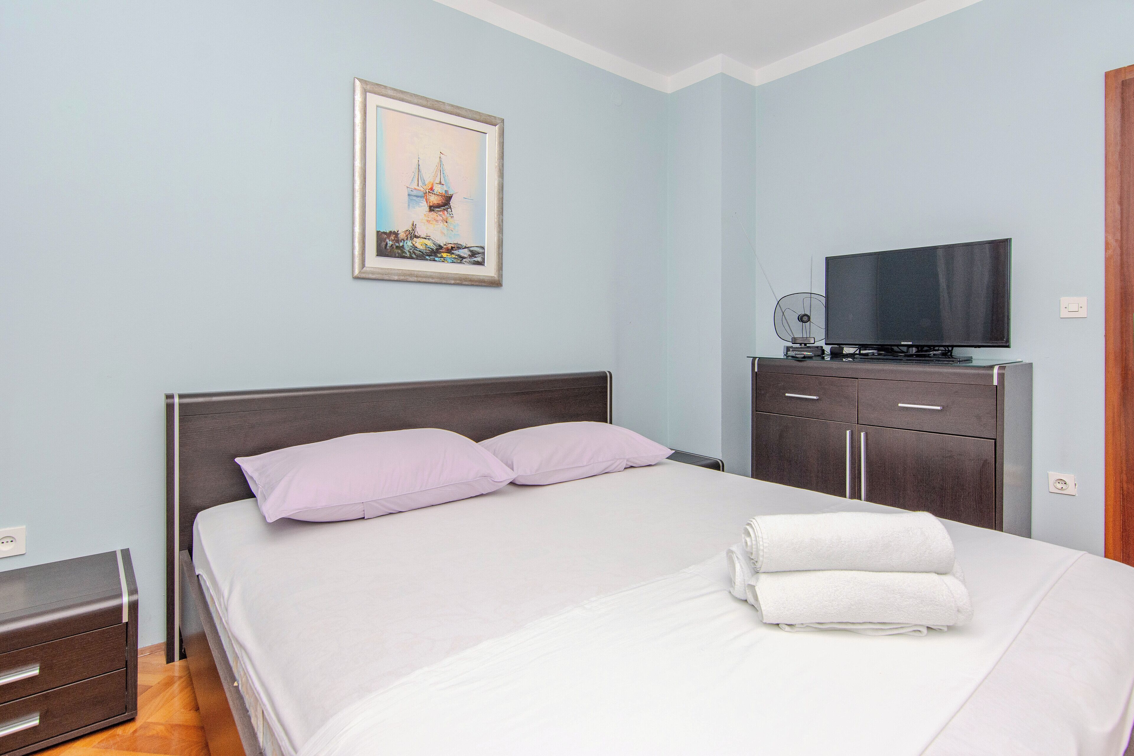 Una cama bien hecha con sábanas blancas y dos almohadas, un televisor sobre un mueble bajo, un aparador de madera y un cuadro en la pared.