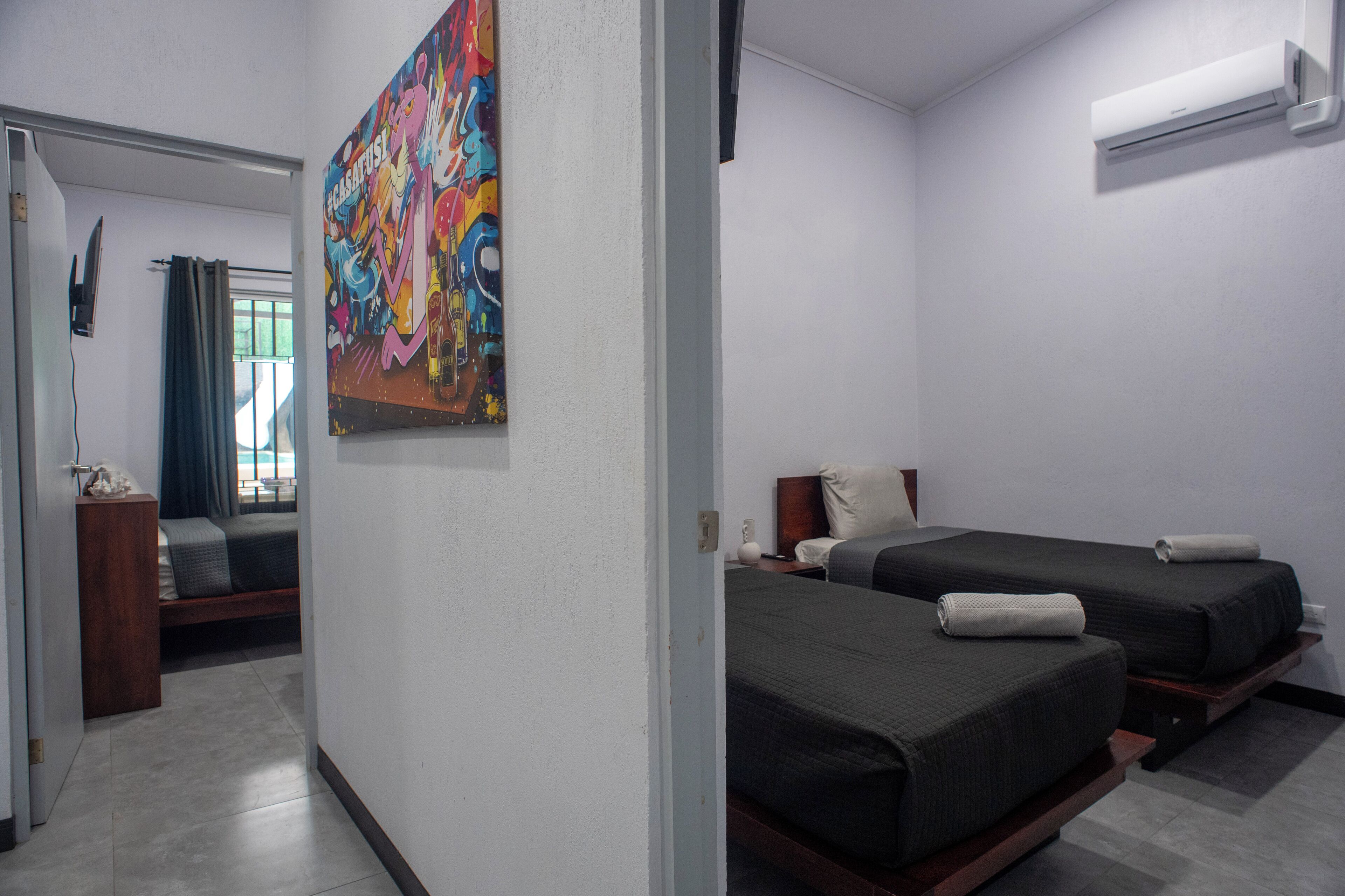 A hotel room with two beds, a colorful abstract painting, a window with curtains, and an air conditioning unit.