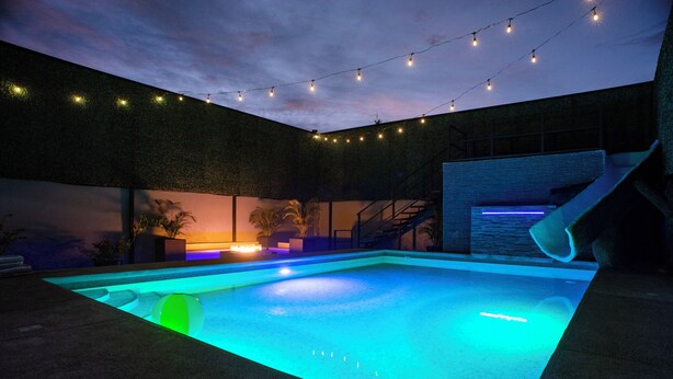 An outdoor swimming pool with a blue-lit edge, string lights, and a staircase leading to a covered area. - Party Villa with Pool, Walk to Clubs (Jacó)