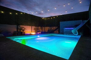 Une piscine extérieure dont les bords sont illuminés en bleu, agrémentée de guirlandes lumineuses et d’un escalier menant à un espace abrité.