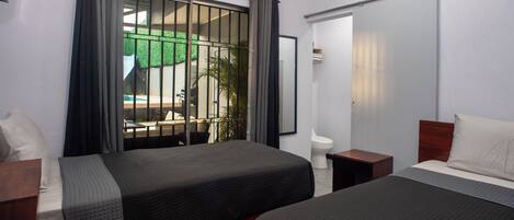 A hotel room with two beds, a window with curtains, an air conditioning unit, and a view of a pool and greenery outside.