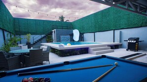 A pool table with billiard balls, a pool cue, and a small pool table in a covered outdoor area with a green wall and a hot tub. - Party Villa with Pool, Walk to Clubs (Jacó)