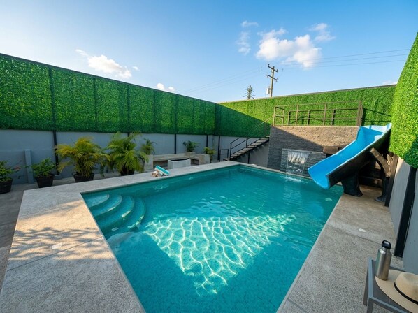 Une piscine avec un toboggan, entourée d’un mur végétal et de plantes en pot.