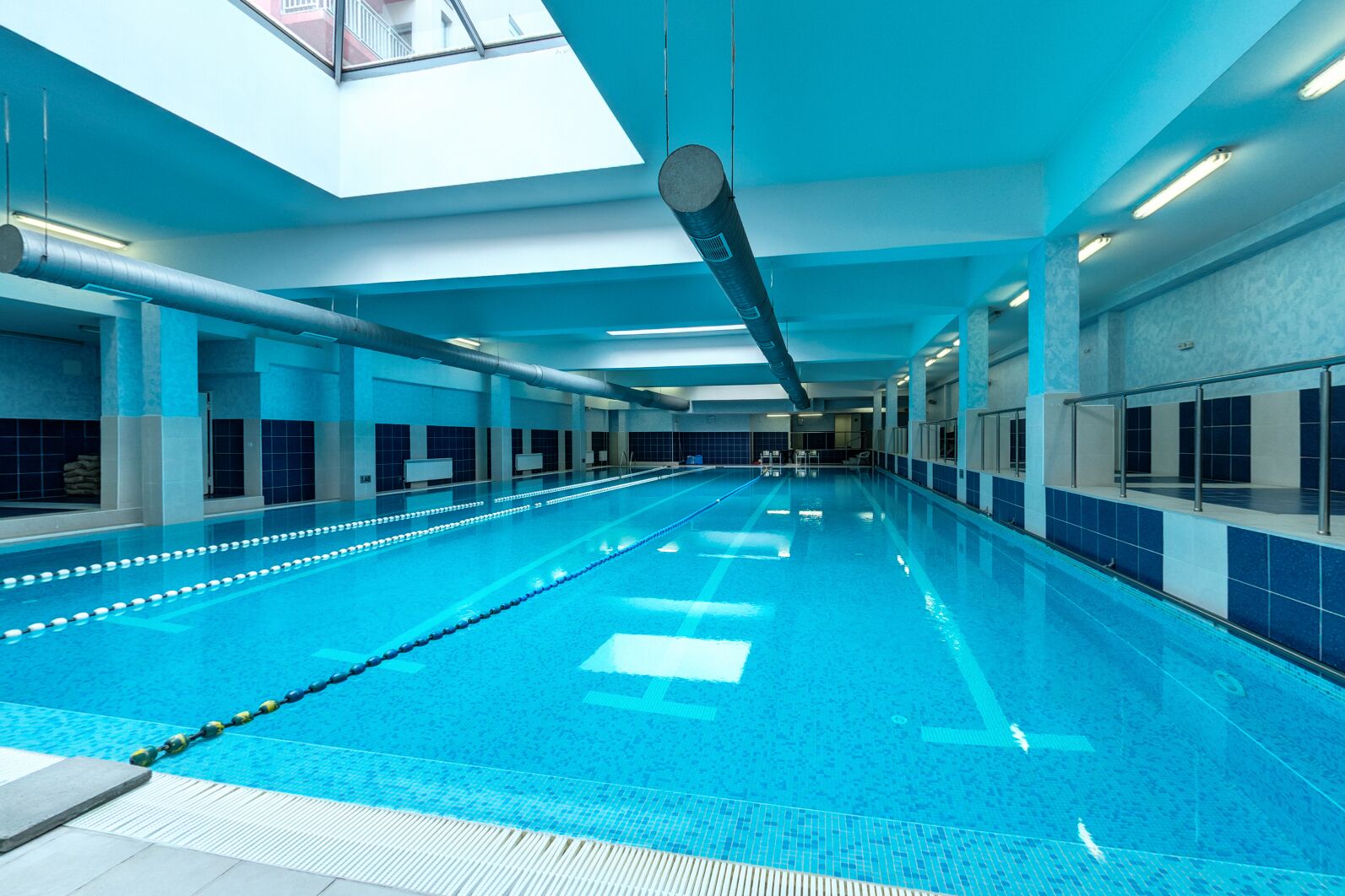 Una amplia piscina interior con carriles delimitados, un techo alto con tragaluces y una pared de azulejos a un lado.