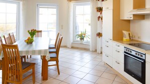 Zona pranzo con tavolo imbandito per quattro, sedie in legno e un vaso di fiori. Si nota una cucina con fornello e pensili.