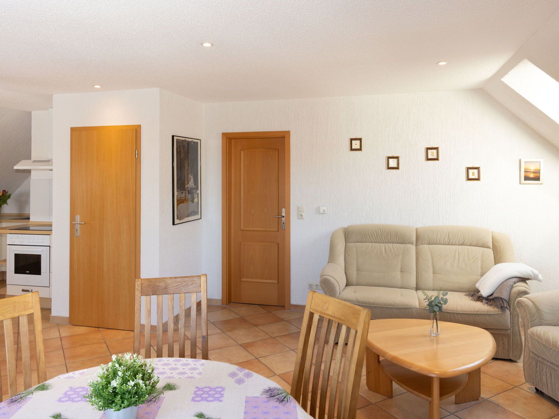 A living room with a beige sofa, wooden chairs, a round table with a vase of flowers, and a kitchen in the background.
