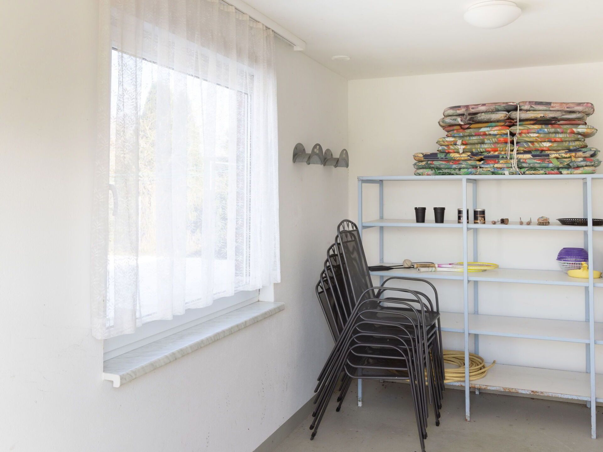 A room with a window, a metal chair, a metal shelf with folded blankets, and a small shelf with various items.