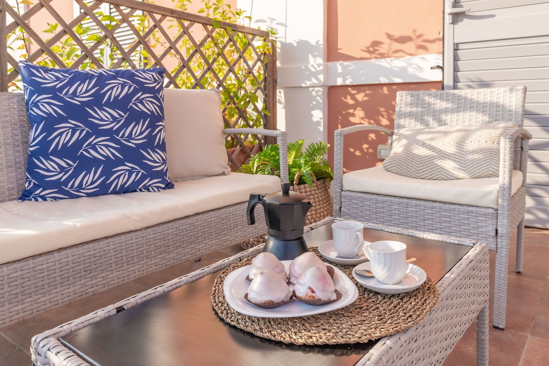 Un canapé en osier agrémenté d’un coussin bleu, d’un autre coussin blanc et d’une table basse sur laquelle se trouve un plateau de chocolats emballés dans du papier blanc.