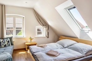 Quarto com teto inclinado, uma cama com almofadas, uma mesa de cabeceira com um vaso e uma janela com cortinas.
