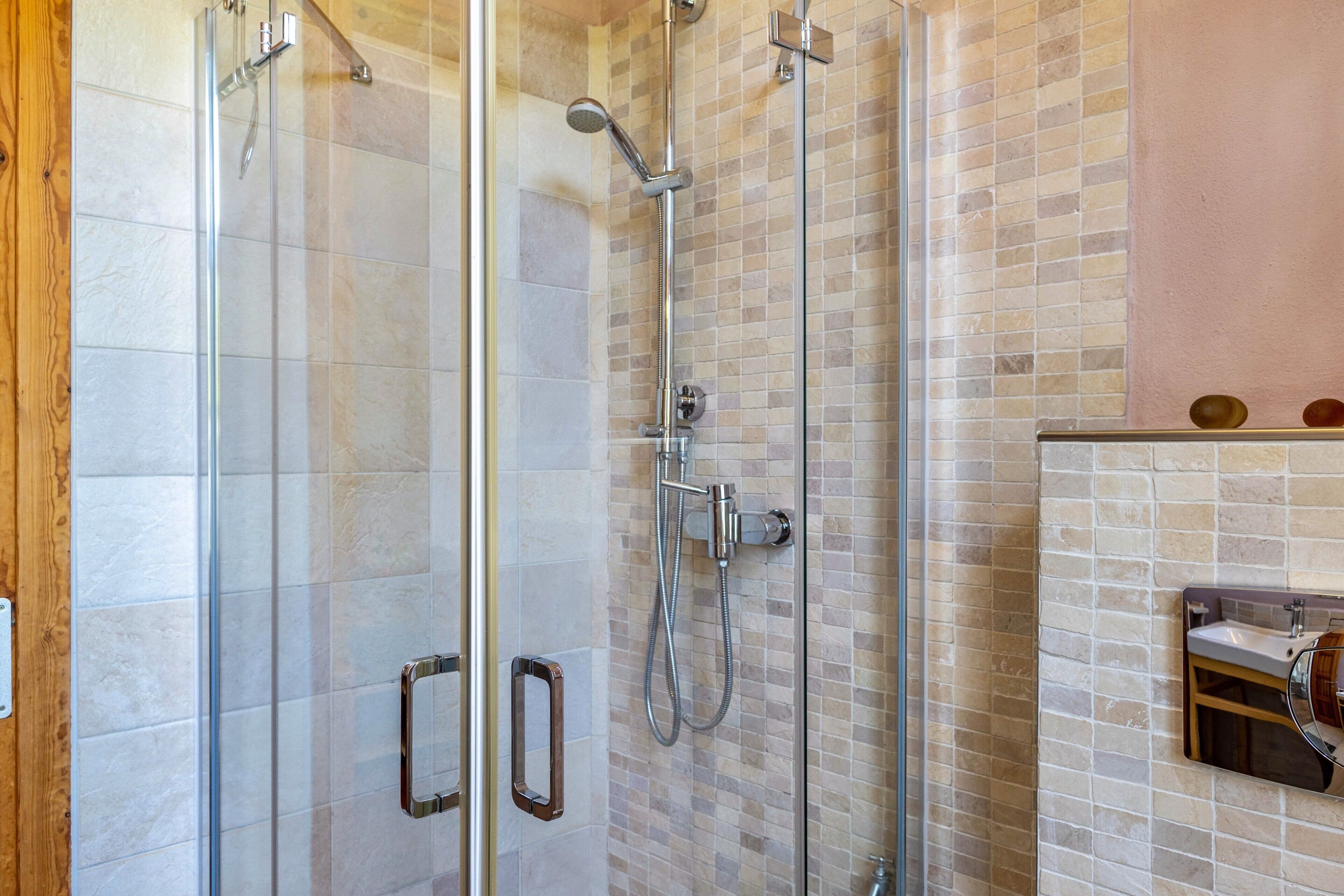 Una ducha con mampara de vidrio, pared con azulejos y un lavamanos pequeño con espejo.