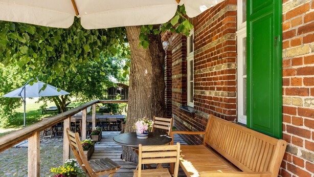 Un patio al aire libre con mobiliario de madera, un muro de ladrillo y un árbol grande que da sombra.