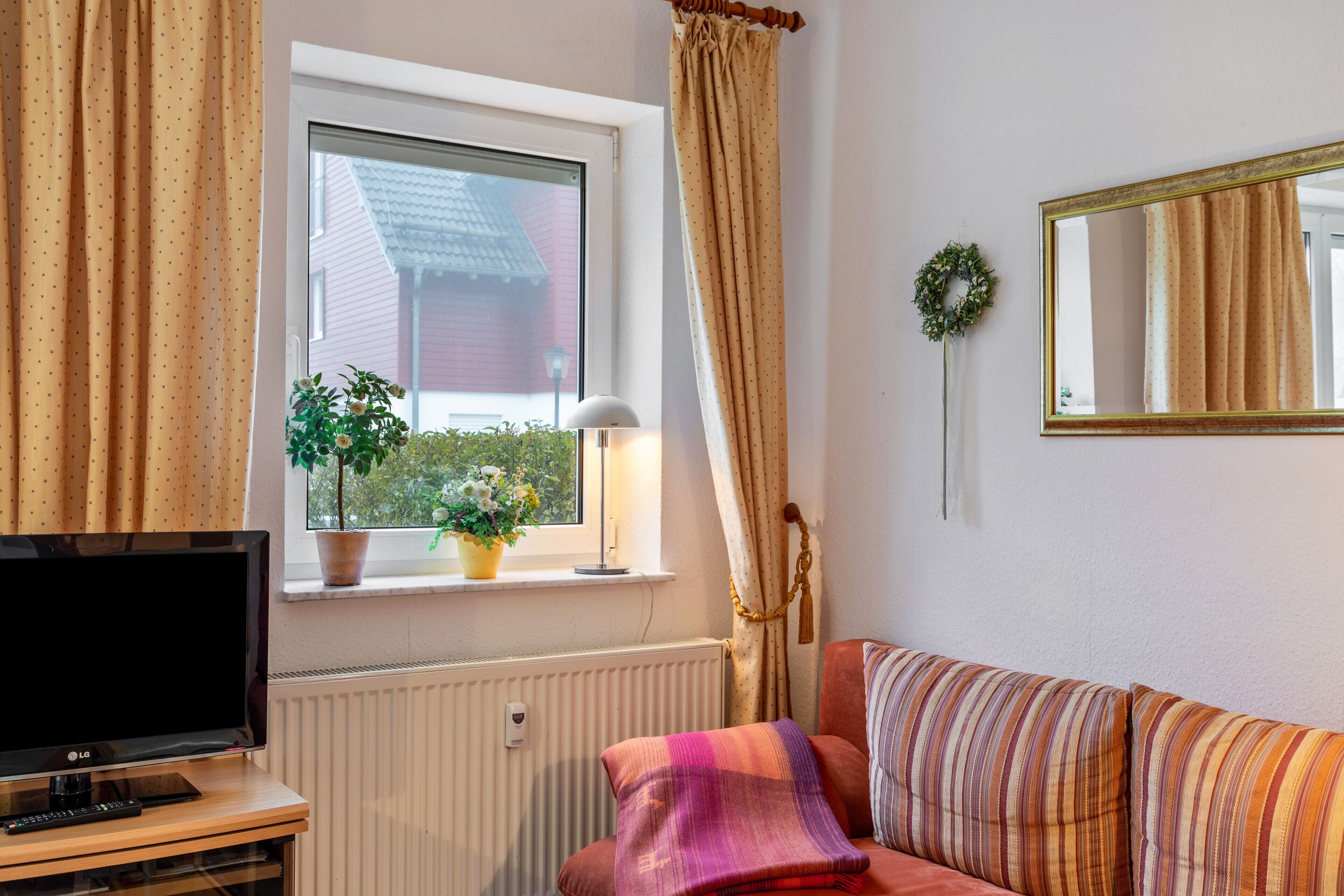 Une pièce avec un téléviseur posé sur un meuble en bois, une fenêtre avec des rideaux, une plante en pot, un canapé avec des coussins à rayures et un miroir accroché au mur.