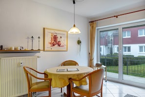 Un espace repas comprenant une table, des chaises, un luminaire suspendu, un radiateur, un tableau encadré et une vue à travers une porte vitrée.
