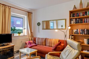 Un salon comprenant un canapé, un fauteuil, un téléviseur, une bibliothèque et une fenêtre avec des rideaux.