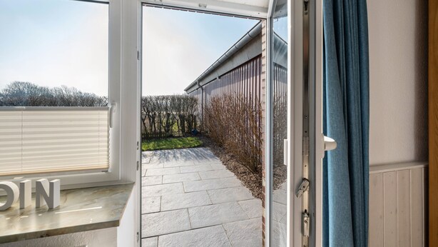 Une porte-fenêtre coulissante donne sur un patio dont le sol est recouvert de pierres, bordé d’une clôture en bois, et qui offre une vue sur les arbres.