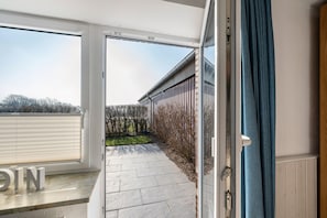 An open sliding glass door leading to a patio with stone flooring, a wooden fence, and a view of trees. - Apartment 'Ferienwohnung - Groß' with Shared Garden and Wi-Fi (Dagebüll)