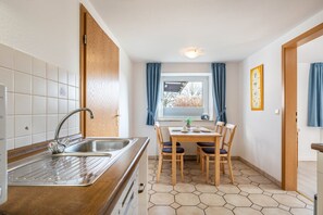 Una cocina con un lavamanos, armarios de madera, una mesa de comedor con sillas, un ventanal con cortinas y un radiador.