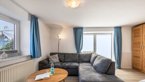 Un salon moderne comprenant un canapé d’angle, une table basse en bois, un radiateur, une fenêtre avec des rideaux et une armoire.
