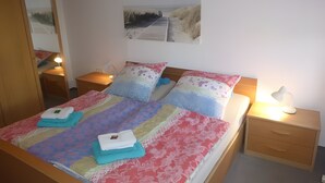 En pænt redt seng med et farverigt sengetæppe, to puder, en sengetøjslampe og et natbord med lampe.