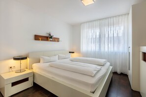 Une chambre d’hôtel moderne, dotée d’un grand lit, de tables de chevet, d’une petite table, d’une étagère murale agrémentée d’une plante et d’une grande fenêtre avec des rideaux légers.